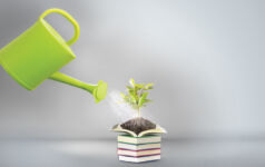 A watering can pouring water over a pile of books with one open at the top and a plant growing out of it